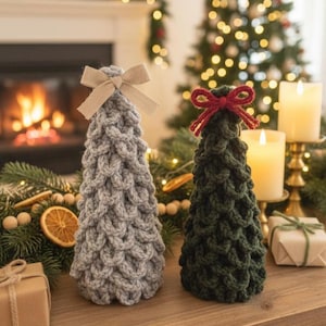 Puede incluir: Dos árboles de Navidad de ganchillo hechos a mano, uno gris y otro verde oscuro, están sobre una superficie de madera. Cada árbol está rematado con un lazo. El fondo incluye una chimenea, un árbol de Navidad, velas y regalos envueltos.