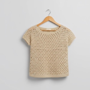May include: A beige, short-sleeved crochet top hangs on a wooden hanger. The top features a round neckline and a loose, open weave design. The neutral color and simple design make it a versatile garment.