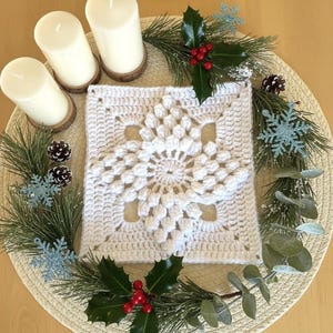 Peut inclure: Une décoration de table de Noël avec trois bougies blanches sur des socles en bois, un napperon en forme de flocon de neige crocheté blanc et une couronne de pin, de houx et de flocons de neige décoratifs. L'ensemble est posé sur un dessous de plat tressé.