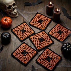 May include: A collection of six orange and black crocheted Halloween coasters. Each coaster features a square design with a black center and border. The coasters are arranged on a wooden surface with Halloween decorations, including pumpkins, candles, and a skull.