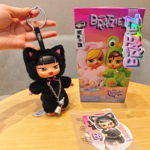 Puede incluir: Un llavero de muñeca Bratz negro con orejas de gato, una cadena plateada y un dije de biberón. La muñeca está junto a una caja de Bratz con muñecas con disfraces de conejito y rana. También es visible una pegatina con una muñeca Bratz de gato.