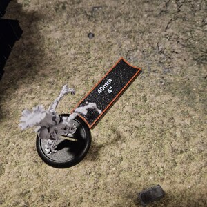 Op de afbeelding: Een zwart-witte miniatuurfiguur van een skeletwezentje met een opgeheven arm, staand op een zwarte basis met een rode rand. De figuur staat naast een zwart-witte liniaal met een oranje rand, gemarkeerd met "40mm 4".