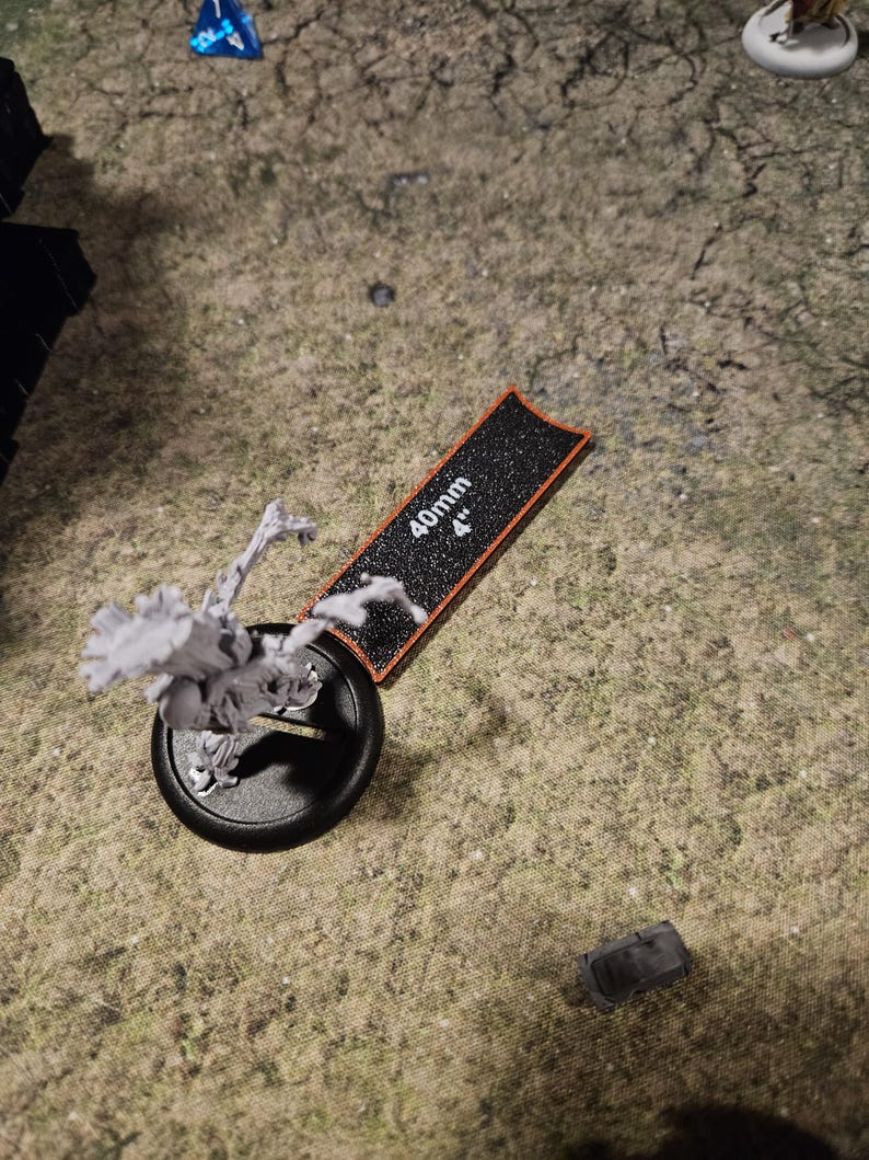 Op de afbeelding: Een zwart-witte miniatuurfiguur van een skeletwezentje met een opgeheven arm, staand op een zwarte basis met een rode rand. De figuur staat naast een zwart-witte liniaal met een oranje rand, gemarkeerd met "40mm 4".