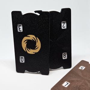 Pode incluir: Cartas de baralho pretas e marrons com números brancos. As cartas pretas têm um design circular dourado. Uma carta marrom está virada para cima, exibindo os números 7 e 5. As cartas pretas mostram os números 0, 2, 3 e 8.