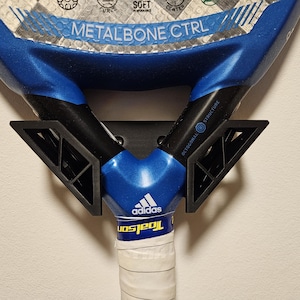 May include: A blue and black paddle tennis racket with a white grip and the text "METALBONE CTRL" and "adidas" on the racket. The racket is mounted on a wall with a black bracket.