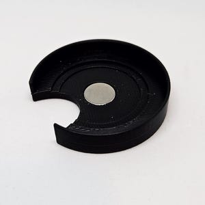 May include: A black, semi-circular 3D-printed object with a recessed center and a silver-colored circular magnet. The object has a curved outer edge and a notch cut out of one side. The object is sitting on a white surface.