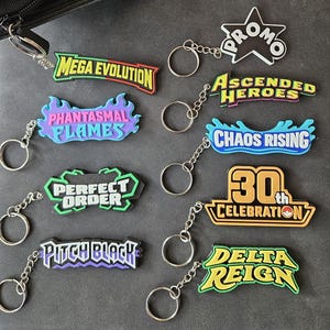 May include: A collection of ten rubber keychains featuring various text designs including Mega Evolution, Phantasmal Flames, Perfect Order, Pitch Black, Promo, Ascended Heroes, Chaos Rising, 30th Celebration, and Delta Reign, each with a metal ring.