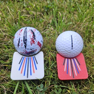 Peut inclure: Deux balles de golf reposent sur des marqueurs colorés dans l'herbe. L'une a un motif éclaboussé, l'autre une ligne noire. Les marqueurs sont blancs et rouges, avec des lignes bleues, rouges et noires.