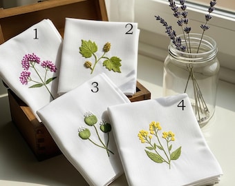 May include: Four white embroidered handkerchiefs with floral designs. The embroidery features purple, green, and yellow flowers. The handkerchiefs are arranged in a wooden box, with a glass jar of lavender in the background.