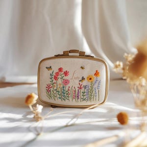 May include: A rectangular jewellery box with a cream-coloured fabric top featuring colourful embroidered wildflowers, a bee, and a butterfly. The box has a metallic gold-toned frame and a small handle on top. The name "Luna" is embroidered in cursive.