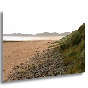 May include: A scenic landscape photograph of a beach with a sandy shore and a rocky foreground. The beach is bordered by grassy dunes on the right and distant hills under an overcast sky. The image captures a natural coastal scene.