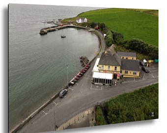 May include: Aerial view of a coastal scene featuring a harbor, a restaurant with the name "Keating's", and a winding road. The water is a dark gray-green, and the land is covered in green grass. A few cars are parked near the restaurant.
