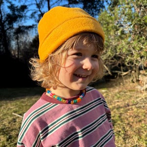 Puede incluir: Un niño con un gorro amarillo mostaza, una camisa de manga larga a rayas rosas y blancas y un collar de cuentas de colores. El fondo presenta árboles y luz solar.