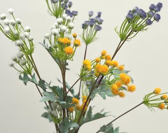 Puede incluir: Un jarrón de cerámica blanca con borde azul contiene un ramo de flores artificiales. Las flores incluyen racimos blancos, amarillos y morados sobre tallos y hojas verdes. El fondo es blanco liso.