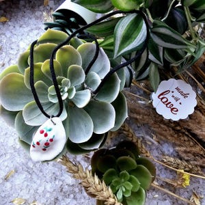 以下が含まれることがあります： 赤と緑のチェリーが付いた白いセラミックペンダントネックレス。ネックレスは黒いコードに付いており、緑の多肉植物の上に置かれています。タグには「愛情込めて作られました」と書かれています。