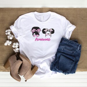 May include: White t-shirt with a graphic of two stylized women's faces with sunglasses and bows. The word "Awareness" is written in pink script below the faces. The shirt is paired with blue jeans and tan ankle boots.