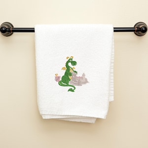 Könnte beinhalten: Weißes Badetuch mit einem gestickten Design eines grünen Drachen, der eine Sandburg baut. Ein rosa Eimer befindet sich daneben. Das Handtuch hängt an einer dunkelbraunen Handtuchstange.