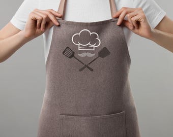 Könnte beinhalten: Eine braune Schürze mit einem Design aus Kochmütze, Schnurrbart und gekreuzten Pfannenwendern. Die Schürze hat eine große Vordertasche und beige Träger. Das Design ist in Weiß und Schwarz gehalten. Eine Person mit weißem T-Shirt hält die Schürze.