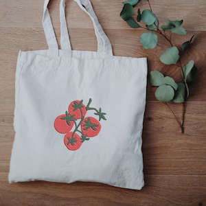 May include: A white canvas tote bag featuring an embroidered design of four red tomatoes with green stems and leaves. The bag has two handles and is made of a light-colored fabric. The design is centered on the front of the bag.