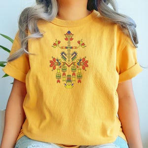 Könnte beinhalten: Senfgelbes T-Shirt mit einem farbenfrohen, geometrischen Kreuzdesign. Das Kreuz besteht aus verschiedenen Formen und Farben, darunter Rot, Grün, Blau und Gelb. Das Shirt hat kurze Ärmel und einen Rundhalsausschnitt.