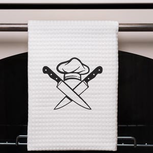 Op de afbeelding: Witte keukendoek met een zwart ontwerp van een koksmuts en gekruiste messen.