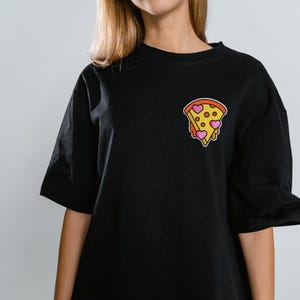 May include: Black t-shirt featuring a pizza slice graphic with heart-shaped pepperoni. The pizza slice has a yellow crust, red sauce, and pink heart-shaped pepperoni. The t-shirt is a casual, everyday wear item.