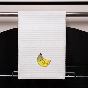 May include: White waffle-weave kitchen towel with an embroidered banana design. The banana features yellow, green, and brown colors. The towel is hanging on a metal bar, likely in a kitchen setting.