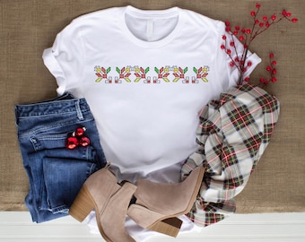 May include: White t-shirt with a colorful floral cross-stitch design across the chest. The shirt is paired with blue jeans, tan ankle boots, and a plaid scarf. Red Christmas ornaments and a sprig of red berries complete the ensemble.