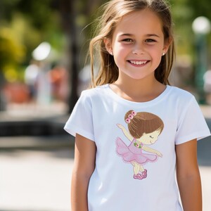 May include: A white t-shirt with an embroidered design of a ballerina girl in a pink tutu. The ballerina is smiling and has brown hair with pink flowers.
