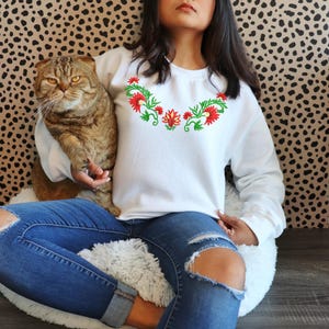 May include: White sweatshirt with a floral design in red and green. The crew neck sweatshirt is paired with blue jeans. A cat is being held in the sweatshirt.