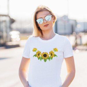 Pode incluir: Camiseta branca com um design colorido de girassóis no peito. O gráfico inclui girassóis amarelos com centros marrons e folhas verdes. A camiseta é um estilo clássico de gola redonda.