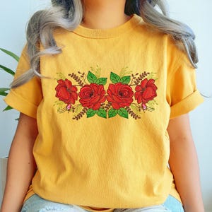 May include: Mustard yellow t-shirt featuring an embroidered floral design. The design includes four red roses with green leaves and small sprigs. The shirt has short sleeves and a relaxed fit, perfect for casual wear.