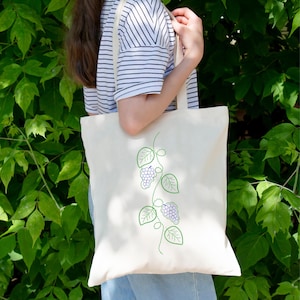 May include: Cream-colored tote bag with a green embroidered grape vine design. The design features clusters of purple grapes and green leaves. The bag has a simple, minimalist style and is carried by a person wearing a striped shirt.