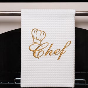 Op de afbeelding: Een witte keukendoek met een gouden geborduurde koksmuts en het woord "Chef" in een cursief schrift.