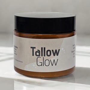 Op de afbeelding: Een bruine pot met een zwart deksel, met het opschrift "Tallow Glow" in een vet, schreefloos lettertype. De pot heeft een wit etiket met extra tekst en sterrenafbeeldingen. Het product is waarschijnlijk een huidcrème of balsem.