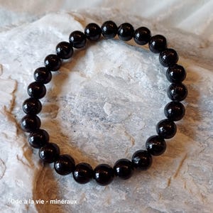 May include: A black beaded bracelet made of round, polished stones. The bracelet is on a light grey, textured surface.