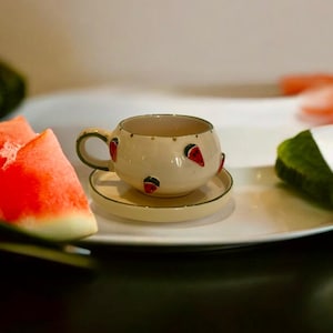 Può includere: Un set tazza e piattino color crema con un design a forma di anguria. La tazza ha una forma arrotondata con un piccolo manico ed è decorata con fette di anguria rosse. Il piattino è in tinta crema con un bordo verde.