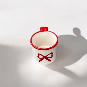 Könnte beinhalten: Eine kleine, weiße Keramik-Tasse mit rotem Schleifenmuster und rotem Rand. Der Griff ist ebenfalls rot. Die Tasse ist zylindrisch und scheint leer zu sein. Die Tasse steht auf einer weißen Oberfläche.