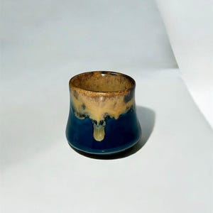 May include: A small, handmade ceramic cup with a unique design. The cup features a dark blue base, transitioning to a tan and brown rim. A single drip of tan glaze runs down the side. This handcrafted piece is perfect for espresso or small beverages.