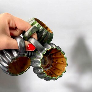 May include: Three ceramic mugs with a ribbed, pumpkin-like design. The mugs are primarily silver with green and brown accents. The handles are thick and ring-shaped. The interior of the mugs is a warm brown color.