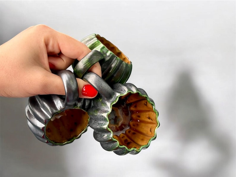 May include: Three ceramic mugs with a ribbed, pumpkin-like design. The mugs are primarily silver with green and brown accents. The handles are thick and ring-shaped. The interior of the mugs is a warm brown color.