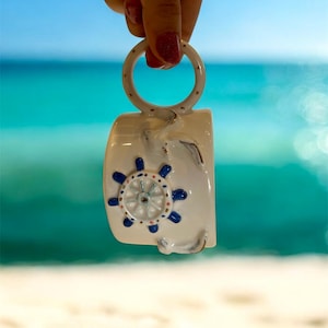 Op de afbeelding: Een witte keramische mok met een nautisch thema, met een scheepswielontwerp in blauw en wit. De mok heeft een cirkelvormige handgreep en wordt vastgehouden tegen een achtergrond van een blauwe oceaan en een zandstrand.