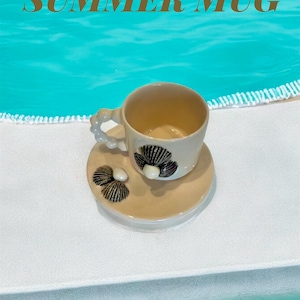 Op de afbeelding: Een beige keramische mok en schotel set met een schelpmotief. De mok heeft een uniek handvat en staat op een wit oppervlak met de tekst "SUMMER MUG" erboven. De achtergrond is turquoise blauw.