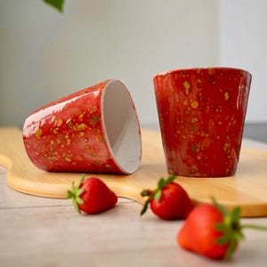 Può includere: Due tazze in ceramica rossa con un design maculato, una in piedi e una di lato, sono su un tagliere di legno. Tre fragole fresche sono sparse nelle vicinanze. Le tazze hanno un interno bianco.