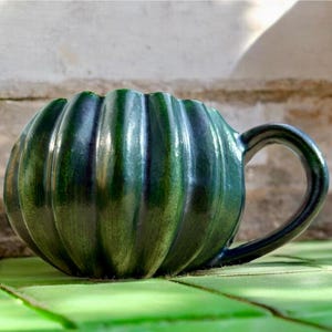 May include: A handmade, green ceramic mug with a ribbed pumpkin shape. The mug has a dark handle and a glossy finish. The mug is sitting on a green surface. This unique mug is perfect for coffee, tea, or any beverage.