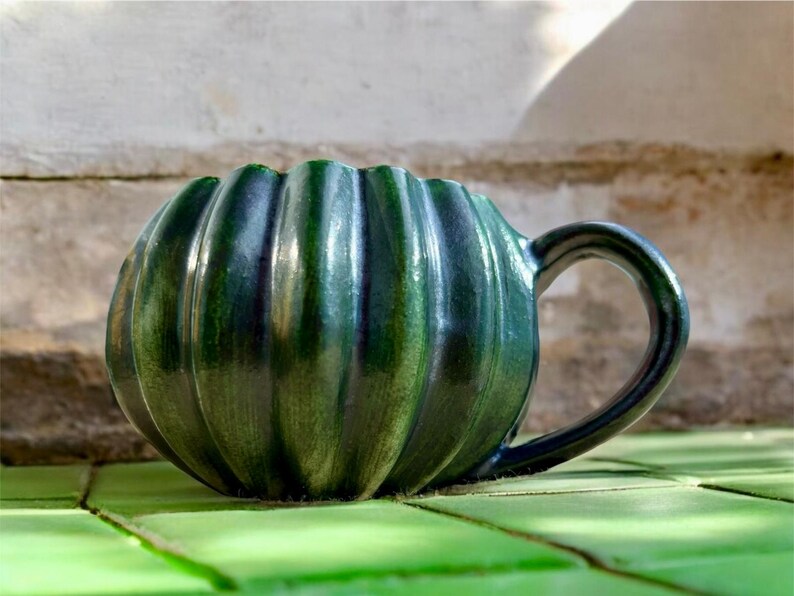 May include: A handmade, green ceramic mug with a ribbed pumpkin shape. The mug has a dark handle and a glossy finish. The mug is sitting on a green surface. This unique mug is perfect for coffee, tea, or any beverage.