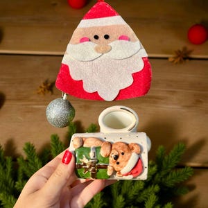 May include: A felt Santa Claus ornament with a red hat and white beard hangs above a small ceramic Christmas decoration. The ceramic piece features a bear in a Santa suit and a green gift box.
