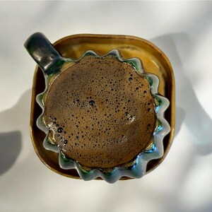 May include: A ceramic coffee cup with a dark brown beverage, resting on a matching saucer. The cup has a unique, wavy rim with green and blue accents. The handle is dark green. The saucer is a light brown color.
