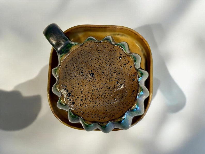 May include: A ceramic coffee cup with a dark brown beverage, resting on a matching saucer. The cup has a unique, wavy rim with green and blue accents. The handle is dark green. The saucer is a light brown color.