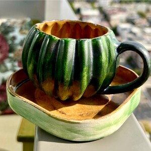 May include: A ceramic teacup and saucer set. The teacup is shaped like a green pumpkin with a black handle. The saucer is square with a light green rim and a brown interior. The set is on a table with a blurred background.